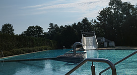 Zu sehen ist das Schwimmbecken mit einer breiten Wasserrutsche. Im Hintergrund ist der blaue Himmel und ein Waldstück zu sehen.