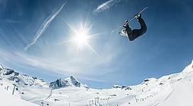 Ein Snowboarder springt in die Luft über einem schneebedeckten Hang. Der Himmel ist klar und sonnig mit einigen Wolken.