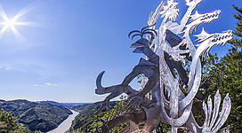 Predigtstuhl in Waldhausen im Strudengau: Eine detaillierte Skulptur eines fantastischen, gefährlich aussehenden Wesens, das auf einem felsigen Vorsprung thront. Das Wesen hat einen mächtigen, muskulösen Körper mit langen, gekrümmten Klauen und Hörnern. Es scheint einen dramatischen Ausdruck und eine bedrohliche Haltung einzunehmen, während es den Blick in die Ferne richtet. Die Skulptur ist aus metallischem Material gefertigt und weist eine komplexe, ornamentale Gestaltung auf. Sie befindet sich an einem exponierten Standort, von dem aus sich eine malerische Landschaft mit bewaldeten Bergen und einem Fluss erstreckt.