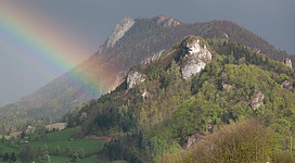 Ein Regenbogen spannt sich über einen grünen Hügel mit felsigen Gipfeln. Der Himmel ist bewölkt, was eine stimmungsvolle Atmosphäre schafft.