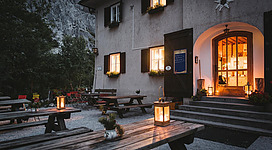 Ein gemütliches Gasthaus mit beleuchteten Fenstern und warmem Licht. Rund um die Holztische sind Laternen platziert, die eine einladende Atmosphäre schaffen.