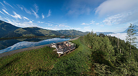Eine malerische Landschaft mit sanften Hügeln und einem ruhigen See. Im Vordergrund befindet sich eine Aussichtsplattform umgeben von Bäumen.