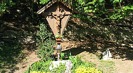 Ein Holzkreuz mit einer Statue von Jesus hängt an einem kleinen Schrein. Umgeben von Blumen und grünen Pflanzen im Wald.