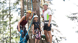 Eine aufregende Klettererfahrung im Wald. Eine Familie steht auf einer Plattform mit Sicherheitsausrüstung und lächelt in die Kamera.