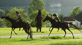 Atelier Fahrner - Skulpturenpark in Waldkirchen am Wesen: Eine Gruppe von Reitskulpturen aus Metall auf einer grünen Wiese, umgeben von Bäumen und einem Gebäude in der Ferne. Die detaillierten Figuren zeigen Pferde und Reiter in dynamischen Posen. Die Skulpturen wirken robust und verleihen der Landschaft eine künstlerische Note.