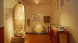 Ein Ausstellungsraum mit historischen Artefakten. Zu sehen sind eine große Steinsäule, eine Tafel und Vitrinen mit weiteren Exponaten.