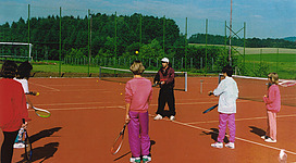 Tennisplatz in St. Aegidi: Mehrere Personen spielen Tennis auf einem Tennisplatz mit roter Aschebahn. Ein Trainer steht in der Mitte und gibt Anweisungen. Die Spieler tragen farbenfrohe Sportkleidung und haben Tennisschläger in den Händen. Hinter dem Platz befindet sich eine Landschaft mit Wald und Wiesen.