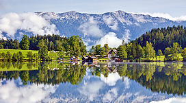 Eine ruhige Landschaft mit einem klaren See und reflektierenden Bergen. In der Nähe sind grüne Wiesen und mehrere kleine Häuser zu sehen.