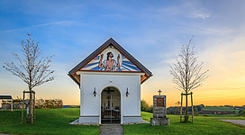 Kalvarienberg - höchste Erhebung mit einzigartigem Fernblick in Haibach ob der Donau: Eine hübsche weiße Kapelle mit einem dreieckigen Giebel und einem Kreuz an der Spitze. An der Frontseite befindet sich ein farbiges, abstraktes Motiv. Umgeben ist die Kapelle von grünen Rasenflächen, vereinzelten Bäumen und einem schönen Sonnenuntergangshimmel.
