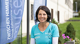 Eine lächelnde Frau steht im Freien vor einem Gebäude. Im Hintergrund sind Fahnen mit einer Werbebotschaft zu sehen.