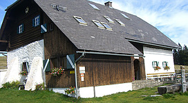 Ein traditionelles Holzhaus mit einem steilen Dach und bunten Fenstern. Das Gebäude steht in einer grünen Wiesenlandschaft unter einem blauen Himmel.