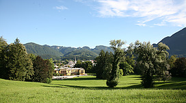 Kaiserpark in Bad Ischl: Das Bild zeigt eine malerische Landschaft mit einem historischen Gebäude, umgeben von grünen Wiesen und Bäumen. Im Hintergrund sind steil aufragende Berge zu sehen, die eine beeindruckende Kulisse bilden.