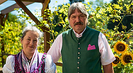 Ein Paar steht vor sonnigen Sonnenblumen und grüner Landschaft. Beide tragen traditionelle Kleidung und lächeln in die Kamera.