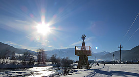 Eine Winterlandschaft mit schneebedecktem Weg und einem Fluss. Im Hintergrund steht ein Aussichtsturm unter einem hellen Sonnenschein.