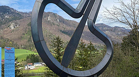 Eine große, moderne Skulptur in Form eines kontrahierten Instruments steht vor einer Berglandschaft. Der Himmel ist klar und es sind Bäume im Vordergrund sichtbar.
