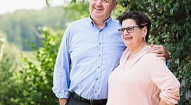 Ein lächelndes Paar steht in einem grünen Garten mit blühenden Pflanzen. Sie wirken entspannt und glücklich in ihrer Umgebung.
