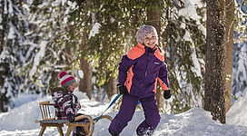 Zwei Kinder spielen im Schnee. Eines zieht einen Schlitten durch einen winterlichen Wald.