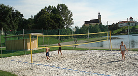 Beach-Volleyballplatz-Badesee in Waldhausen im Strudengau: Eine Strandvolleyballanlage an einem See oder Fluss. Im Vordergrund sieht man einen Sandplatz mit Volleyballnetz, dahinter erstreckt sich eine Parklandschaft mit Bäumen und in der Ferne ist ein Kirchturm zu erkennen.