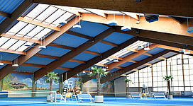 Ein modernes Tenniscenter mit blauen Spielflächen und einer hohen Holzdecke. Palmblätter und große Fenster sorgen für ein helles und einladendes Ambiente.