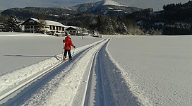 Eine Person in rot zieht mit Langlaufskiern eine Spur durch den frischen Schnee. Im Hintergrund sind schneebedeckte Berge und ein Haus zu sehen.