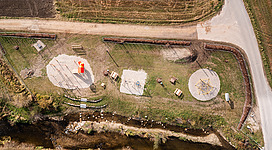 Archi's Natur- Erlebnis- und Generationenplatz in Kirchheim im Innkreis: Eine Fläche aus Gras und Erde, mit strukturierten Elementen wie runden Formen und langen Strukturen, die sich über das Gelände erstrecken. Einige kleinere Objekte sind auf der Fläche verteilt.