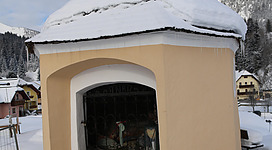 Ein kleiner, runder Pavillon mit einem schneebedeckten Dach steht in einer verschneiten Landschaft. Im Hintergrund sind Bäume und Häuser sichtbar.