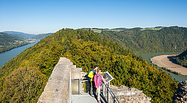 Panoramablick Burgruine Haichenbach in Hofkirchen im Mühlkreis: Eine Aussichtsplattform befindet sich an einem bewaldeten Hang mit einem Blick auf einen Fluss und die umliegenden Berge. Zwei Personen, eine in gelber und eine in rosa Kleidung, befinden sich auf der Plattform und genießen offensichtlich die Aussicht.