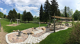 Ein schöner Spielplatz mit Schaukeln und einem Wasserspielbereich. Umgeben von viel Grün und Bäumen bietet er eine einladende Atmosphäre für Kinder.
