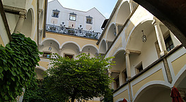 Ein schöner Innenhof mit Säulen und einem grünen Baum. Im Hintergrund ist ein Gebäude mit dekorativen Fenstern und einem klaren Himmel zu sehen.