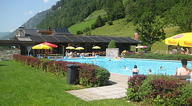 Freibad in den Bergen mit mehreren Badegästen im Wasser. Um das Becken stehen Sonnenschirme, im Hintergrund sind grüne Berge zu sehen.