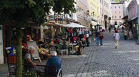 Eine belebte Gasse mit bunten Gebäuden und Geschäften. Einge Bürger sitzen auf einer Bank und genießen die Atmosphäre.