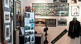 Schauraum der Bergknappenkapelle in Wolfsegg am Hausruck: Ein Raum mit Wänden aus Holz und Regalen, an denen Fotos, historische Dokumente und Uniformen ausgestellt sind. In der Mitte steht ein Podest mit Objekten, die an eine historische Ausstellung erinnern. An den Wänden hängen weitere Bilder und Memorabilien, die einen Einblick in die Vergangenheit geben.