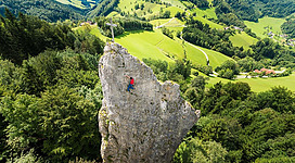 Ein Abenteurer klettert eine große Felsformation inmitten von grünen Hügeln. Im Hintergrund sind weitläufige Landschaften und Bäume sichtbar.