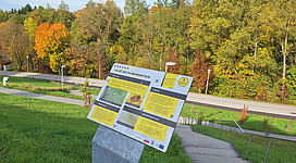 Ein Informationstafel steht an einem Weg mit buntem Blätterdach im Hintergrund. Die Umgebung zeigt einen klaren Himmel und eine ruhige, grüne Landschaft.
