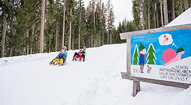 Eine Gruppe von Menschen fährt mit Schlitten einen verschneiten Hang hinunter. Neben dem Weg steht ein bunter Schilder mit einem fröhlichen Bild und einer Aufforderung zum Spaß im Schnee.