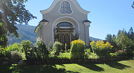 Ein hübsches Gebäude mit einer geschwungenen Dachform, umgeben von üppigem Grün und bunten Blumen. Im Hintergrund sind sanfte Hügel sichtbar unter einem klaren blauen Himmel.