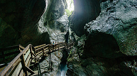 Eine beeindruckende Schlucht mit hohen Felsen und einem Holzsteg. Das Licht strahlt durch eine Öffnung oben und reflektiert im Wasser.