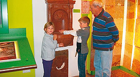 Zwei Kinder und ein älterer Mann stehen neben einem historischen Brunnen in einem Museum. Die Wände sind in lebhaften Farben gestrichen und zeigen verschiedene Materialien.