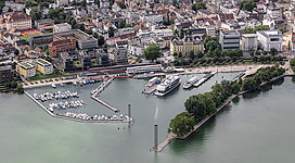 Luftbild vom Hafen Bregenz bei sehr hohem Wasserstand. Links und rechts der Hafeneinfahrt stehen die beiden Leuchttürme. Auf der linken Seite sieht man den Yachthafen (Marina Bregenz), in der Mitte das Hafengelände, das ausschließlich für die Kursschifffahrt bestimmt ist und rechts das Blumenmolo.