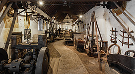 Eine historische Werkstatt mit verschiedenen Maschinen und Werkzeugen. Der Raum hat eine gewölbte Decke und einen gepflasterten Boden.