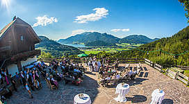 Die Laimeralm in Strobl: Das Bild zeigt eine Terrasse oder ein Außenrestaurant, das von einer malerischen Landschaft mit Bergen und Wäldern umgeben ist. Eine Gruppe von Gästen sitzt an Tischen und genießt offenbar eine Veranstaltung oder ein Fest in dieser wunderschönen Umgebung.