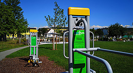 Generationenpark der Begegnung und Bewegung in Perg: In diesem Bild sehe ich einen öffentlichen Sportbereich mit verschiedenen Fitnessgeräten. Die Geräte sind grün und gelb gestaltet und laden zu körperlichen Aktivitäten im Freien ein. Hinter den Sportgeräten befindet sich eine Rasenfläche umgeben von Bäumen, die eine angenehme Umgebung für die Erholung und Bewegung bieten.