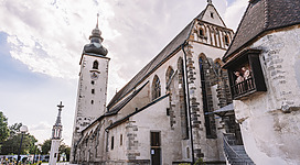 Basilika St. Laurenz in Enns: Eine alte Kirche mit hohem Turm, die sich vor einem bewölkten Himmel erhebt. Die Fassade ist aus Stein gefertigt und weist viele Verzierungen und Bögen auf. Daneben ist ein schlanker Turm mit einer Spitze zu sehen. Im Vordergrund befindet sich ein Platz mit einigen Bänken und Treppen, die zum Haupteingang der Kirche führen.