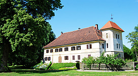 Schloss Mamling in Mining: Ein mehrstöckiges, traditionell gestaltetes Gebäude mit roten Ziegeldächern, umgeben von üppiger Grünfläche und großen Bäumen. Das Gebäude wirkt historisch und gepflegt, mit vielen Fenstern und einem Turm mit Spitzdach. Die Umgebung vermittelt einen idyllischen und ländlichen Charakter.