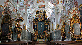 Stiftskirche Garsten in Garsten: Das Bild zeigt eine prachtvolle Kirchenausstattung mit filigran geschnitzten, goldverzierten Altären und Seitenwänden, die mit farbenfrohen Gemälden und Skulpturen dekoriert sind. Das Kircheninnere ist mit imposanten Kronleuchtern und reichen Details ausgestattet, die eine feierliche und ehrfurchtsvolle Atmosphäre vermitteln.