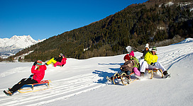 Zwei Kinder spielen im Schnee. Das Mädchen trägt einen roten Schneeanzug, während der Junge in einem grünen Anzug hinterherläuft.