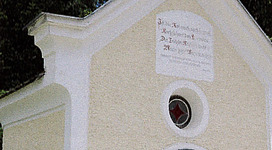 Eine kleine Kapelle mit einer weißen Fassade und einem schmiedeeisernen Tor. Umgeben von hohen Bäumen und blauem Himmel.