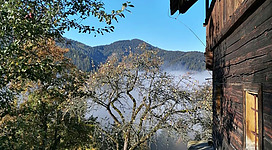 Eine ruhige Berglandschaft mit einem Holzhaus und Nebel in der Ferne. Grüne Wiesen und Bäume umgeben die Szene.