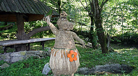 Eine Strohpuppe in Form eines Schweins steht in einer grünen Waldlandschaft. Im Hintergrund sind ein Unterstand und Bäume sichtbar.