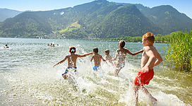 Eine Gruppe von Kindern spielt im Wasser und springt ins Ufer. Im Hintergrund sind Berge und ein blauer Himmel zu sehen.